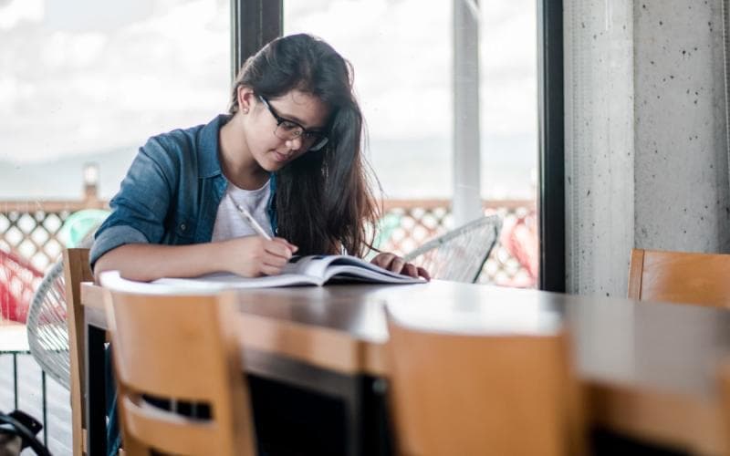 A student writing notes from a textbook at a study table